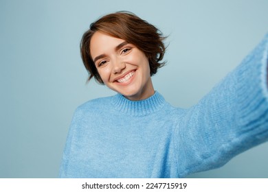 Close up young caucasian woman wear knitted sweater look camera doing selfie shot pov on mobile cell phone isolated on plain pastel light blue cyan background studio portrait. People lifestyle concept