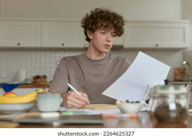 Close up of young curly haired handsome man wearing beige t-shirt sitting at a kitchen table surrounded by comforts of domestic life making notes reviewing papers while having breakfast in kitchen. - Powered by Shutterstock