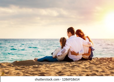 Close Up Of Young Family Sitting Together In Late Afternoon Sun On Beach.Foursome Giving Back Looking At Sea.