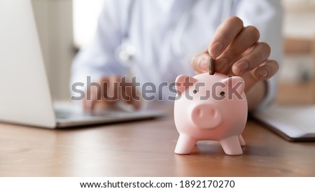 Close up young female general practitioner putting coin in piggy bank, managing hospital budget using laptop accounting applications, medical insurance finances, healthcare money savings concept.