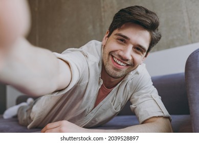 Close up young fun cheerful happy man 20s wearing casual clothes beige shirt pink t-shirt doing selfie shot on mobile phone lying on grey sofa rest indoors at home on weekends. People leisure concept.