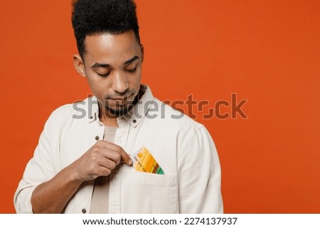 Close up young fun happy man of African American ethnicity wear light shirt casual clothes hold in hand put mock up of credit bank card into pocket isolated on orange red background studio portrait