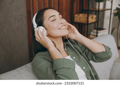 Close up young fun woman of Asian ethnicity wear casual clothes headphones listen music close eyes sit on grey sofa couch stay at home hotel flat rest relax spend free spare time in living room indoor