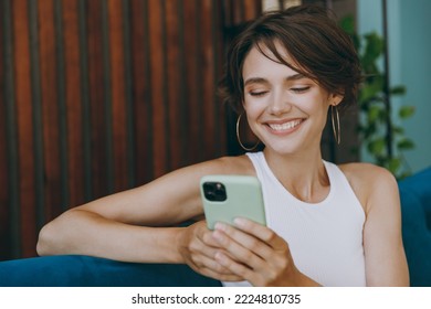 Close up young fun woman wear white tank shirt hold in hand use mobile cell phone sit on blue sofa stay at home hotel flat rest relax spend free spare time in living room indoor People lounge concept - Powered by Shutterstock