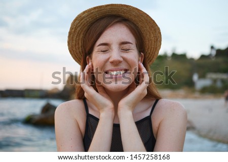 Close up of young ginger cute freckles woman walks on the beach, wears hat, touches cheeks, broadly smiles with closed eyes, looks positive and happy.
