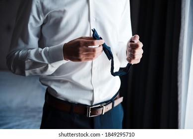 Close Up Of Young Groom In White Shirt And Black Pants Holding In Hands Bow Tie. Man Dressing Up During Morning Time. Wedding Preparation.