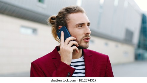 Close Up Of Young Handsome Caucasian Man In Red Jacket Talking On Mobile Phone Outdoors. Serious Good-looking Stylish Male Standing At Street And Speaking On Cellphone. Telephone Conversation.