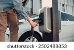 Close up of young handsome male plugging in cable from electric car into charger station outdoor. Man starts charging his electro vehicle in city. EV charger point Electrocar future Zero emission auto