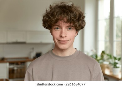 Close up of a young handsome positive curly man with blue eyes with a smiling face wearing casual beige t-shirt looking at camera and standing alone in morning in the modern kitchen at home.