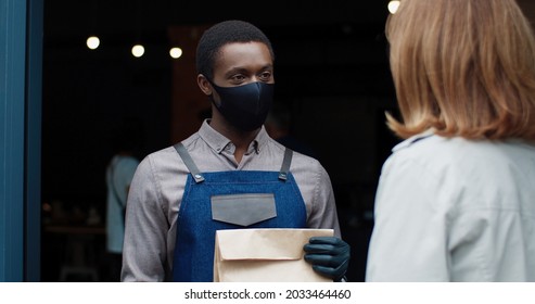 Close Up Of Young Happy African American Handsome Waiter In Black Mask Walk Out Coffee House Giving Order In Paper Bag To Caucasian Female Customer. Restaurant Business During Lockdown. Work Concept