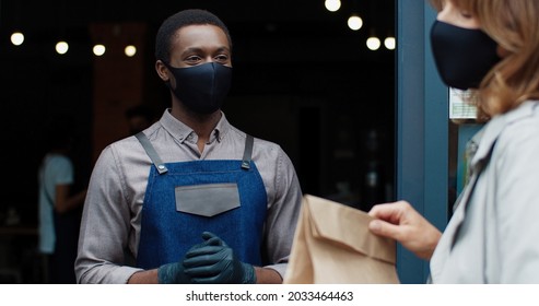 Close Up Of Young Happy African American Handsome Waiter In Black Mask Walk Out Coffee House Giving Order In Paper Bag To Caucasian Female Customer. Restaurant Business During Lockdown. Work Concept