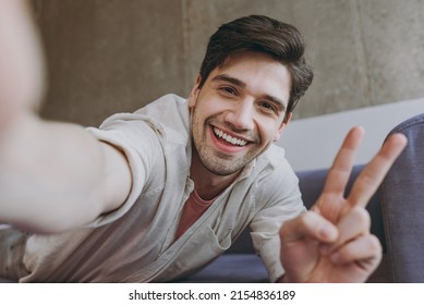 Close up young happy man 20s in casual clothes beige shirt pink t-shirt show victory gesture do selfie shot on mobile phone lying on grey sofa rest indoor at home on weekends People leisure concept.