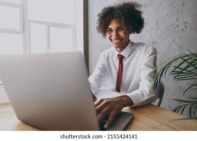 Close up young happy successful african american employee business man 20s in classic shirt tie sit work at white office desk with pc laptop computer typing message inside Achievement career concept