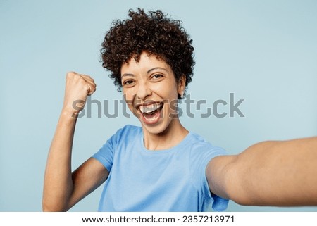Close up young happy woman of African American ethnicity wear t-shirt casual clothes doing selfie shot pov on mobile cell phone do winner gesture isolated on plain pastel light blue cyan background