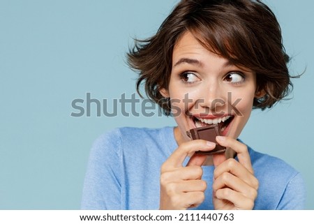 Close up young happy woman in casual sweater hold sweet pink cream donuts biting chocolate bar look camera isolated on plain pastel light blue background studio portrait. People lifestyle food concept