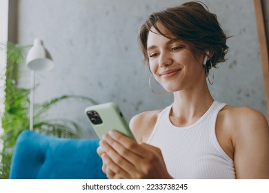 Close up young happy woman wear white tank shirt earphones listen music hold use mobile cell phone sit on blue sofa stay at home flat rest relax spend free spare time in living room indoors grey wall - Powered by Shutterstock