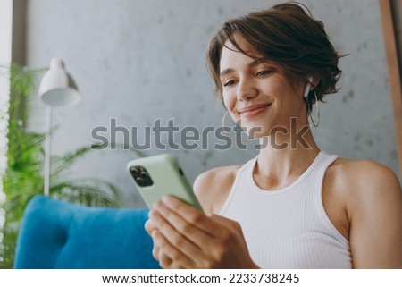 Close up young happy woman wear white tank shirt earphones listen music hold use mobile cell phone sit on blue sofa stay at home flat rest relax spend free spare time in living room indoors grey wall