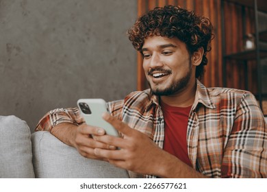 Close up young Indian man wears casual clothes hold in hand use mobile cell phone sits on grey sofa couch stay at home hotel flat rest relax spend free spare time in living room indoor. Lounge concept - Powered by Shutterstock