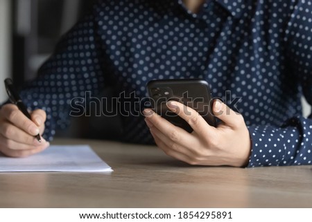 Close up young Indian woman holding smartphone, female student writing notes, preparing exam, studying online, employee intern watching webinar, internet course, searching information, browsing apps