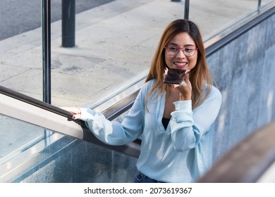 Close up of young latin businesswoman with glasses using smartphone while smiling, going up on metro, subway, airport escalator. Business concept