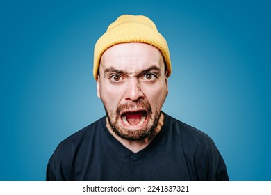 Close up young mad angry displeased guy in yellow cap looking at camera screaming. Young Caucasian angry man yells directly into the camera. Men emotions blue background, Furious man. Stress hysteria. - Powered by Shutterstock