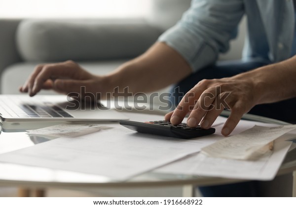 Close up of young male accountant hands make audit\
of expenses calculate charges based on paper documents. Cropped\
shot of man bookkeeper work with bills pay fees taxes online using\
web app on laptop