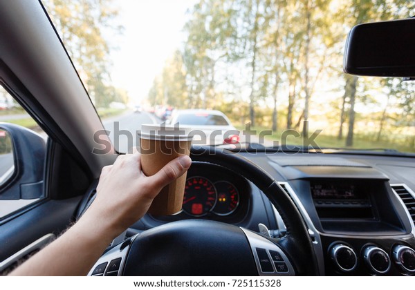 Close up young man car driver drink coffee,
hand holding a Paper Cup of coffee in the background steering the
car dashboard blurry green
background.