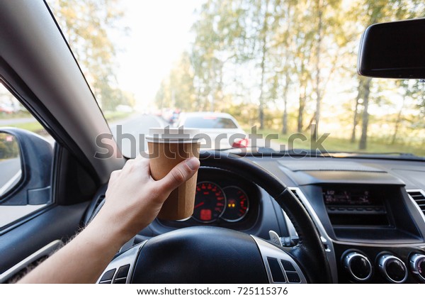 Close up young man car driver drink coffee,\
hand holding a Paper Cup of coffee in the background steering the\
car dashboard blurry green\
background.
