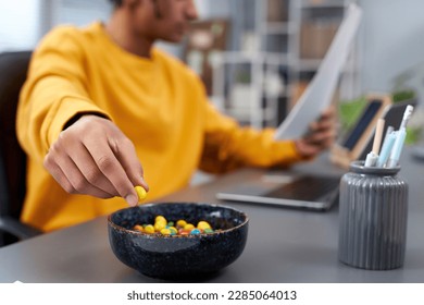 Close up of young man eating snacks while working or studying at home, focus on hand holding colorful candy, copy space - Powered by Shutterstock