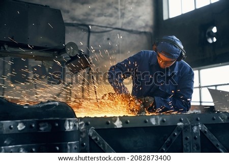 Close up of young man fitter in blue uniform and special headphone welding metal by angle grinder with powerful sparks in smithy. Concept of process working with electrical machine. 
