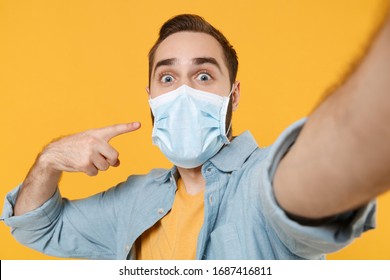 Close Up Of Young Man Isolated On Yellow Background. Epidemic Pandemic Coronavirus 2019-ncov Sars Covid-19 Flu Virus Concept. Doing Selfie Shot On Mobile Phone Point Index Finger On Sterile Face Mask