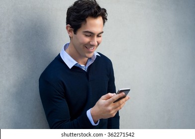 Close Up Of A Young Man Leaning Against A Grey Wall Using Mobile Phone. Portrait Of A Happy Business Man Holding A Smartphone. Man In Casual Typing And Reading A Message On Cell Phone With Copyspace.
