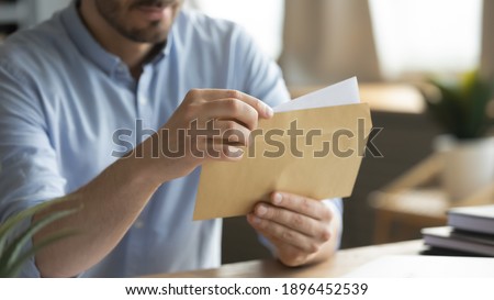 Close up young man opening envelope with paper correspondence. Curious businessman getting paper document by postal service or financial notification at workplace, received notification or invitation.