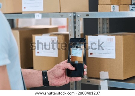 Close up of young man scanning package label with smartphone at order pickup point, managing inventory and delivery workflow. Useful for logistics, ecommerce, fulfillment marketing
