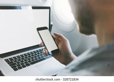 Close Up Of Young Man Sitting At Airplane And Using Smartphone With Blank Screen And Open Laptop, Back View