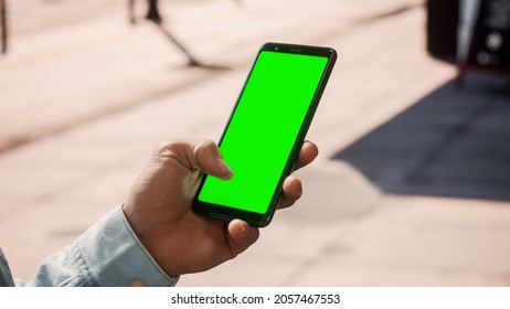 Close Up of a Young Man Using Smartphone with Green Screen Chroma Key Mock Up Display in Vertical Position on a Street During a Sunny Day. He's Tapping and Swiping the Screen. - Powered by Shutterstock