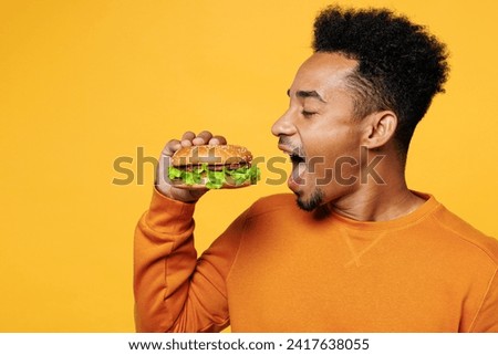 Close up young man wear orange sweatshirt casual clothes hold eat bite burger open mouth isolated on plain yellow background studio portrait Proper nutrition healthy fast food unhealthy choice concept