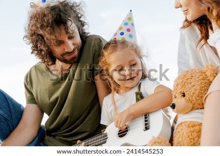 Close up young man and woman sitting on a beach in hats while their cute little daughter playing on ukulele for them while celebrating birthday