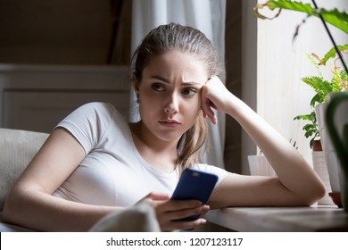 Close up young millennial pensive woman sitting on sofa near windowsill at home holding smartphone waiting message from boyfriend. Irritated female feels annoyed has a problem received unpleasant news