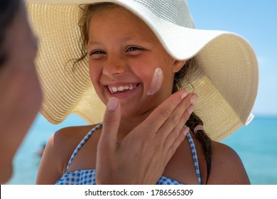 Close Up Of Young Mother Is Applying Protective Sunscreen Or Sunblock Lotion On Her Little Happy Smiling Daughter's Face To Take Care Of Skin On A Seaside Beach During Family Holidays Vacation.
