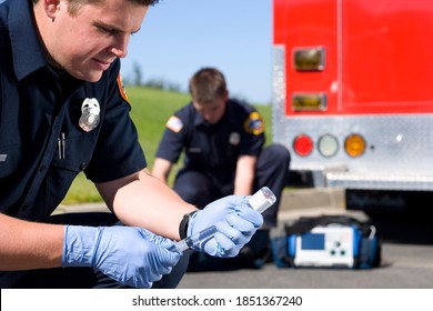 Close up of a young paramedic loading up a syringe while his colleague is taking out other medical equipment from the bag in the background