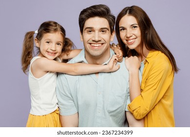 Close up young parents mom dad with child kid girl 6 years old wear blue yellow casual clothes daughter hugs father neck, wife cuddles husband isolated on plain purple background. Family day concept - Shutterstock ID 2367234399