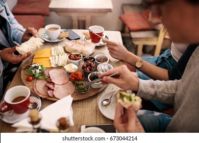 Close Up Of Young People Sitting Around Cafe Table And Eating With Focus On Hands And Food. Group Of Students Having Food In College Canteen.