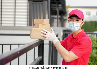 Close Up Young Postal Worker Wear Mask And Glove With Red Uniform Carrying Box And Stand In Front Of House To Give Order To Customer For Contactless Delivery And E-commerce New Normal Concept