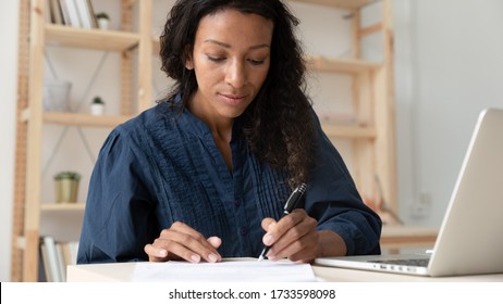 Close up young serious african american businesswoman sign up document at work. Tired black woman employee writing report on paper near modern laptop. Success business deal concept. - Powered by Shutterstock