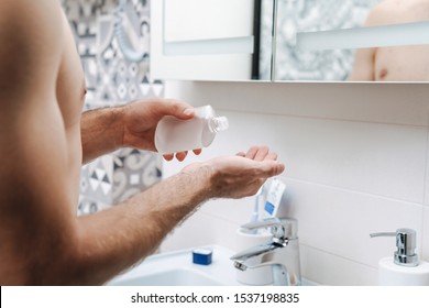 Close up of a young shirtless man applying aftershave lotion while standing at the bathroom mirror