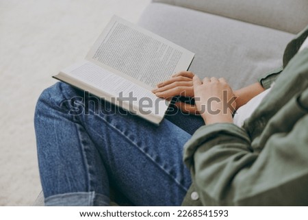 Close up young smart woman wearing green casual clothes reading book novel sits on grey sofa couch stay at home hotel flat rest relax spend free spare time in living room indoor. Hobby leisure concept