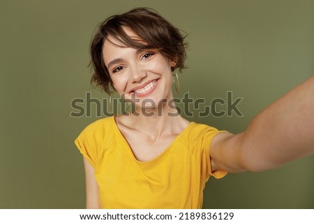 Close up young smiling fun happy woman she 20s wear yellow t-shirt doing selfie shot pov on mobile cell phone isolated on plain olive green khaki background studio portrait. People lifestyle concept
