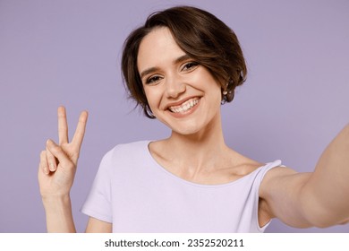 Close up young smiling happy friendly woman 20s with bob haircut wear white t-shirt doing selfie shot on mobile phone show victory v-sign gesture isolated on pastel purple background studio portrait