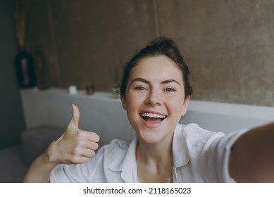 Close up young smiling happy woman in white clothes glasses sit on grey sofa indoors apartment do selfie shot on mobile phone show thumb up gesture Rest on weekend leisure quarantine stay home concept - Powered by Shutterstock